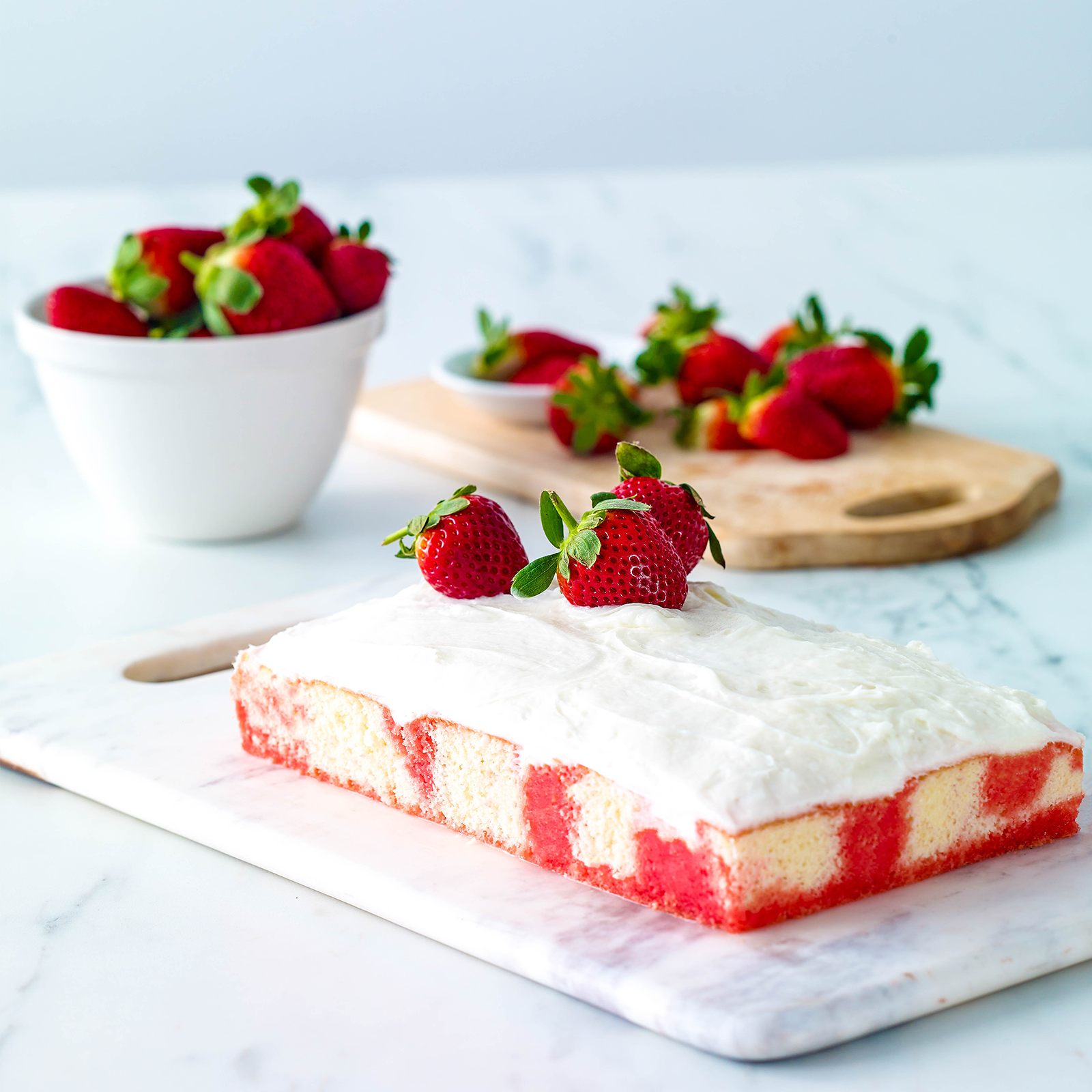 A strawberry poke cake iced with whipped cream reads on a marble cutting board. Fresh strawberries decorated the top of the gluten-free cake. More fresh strawberries are in a bowl and on a wooden board behind the cake.