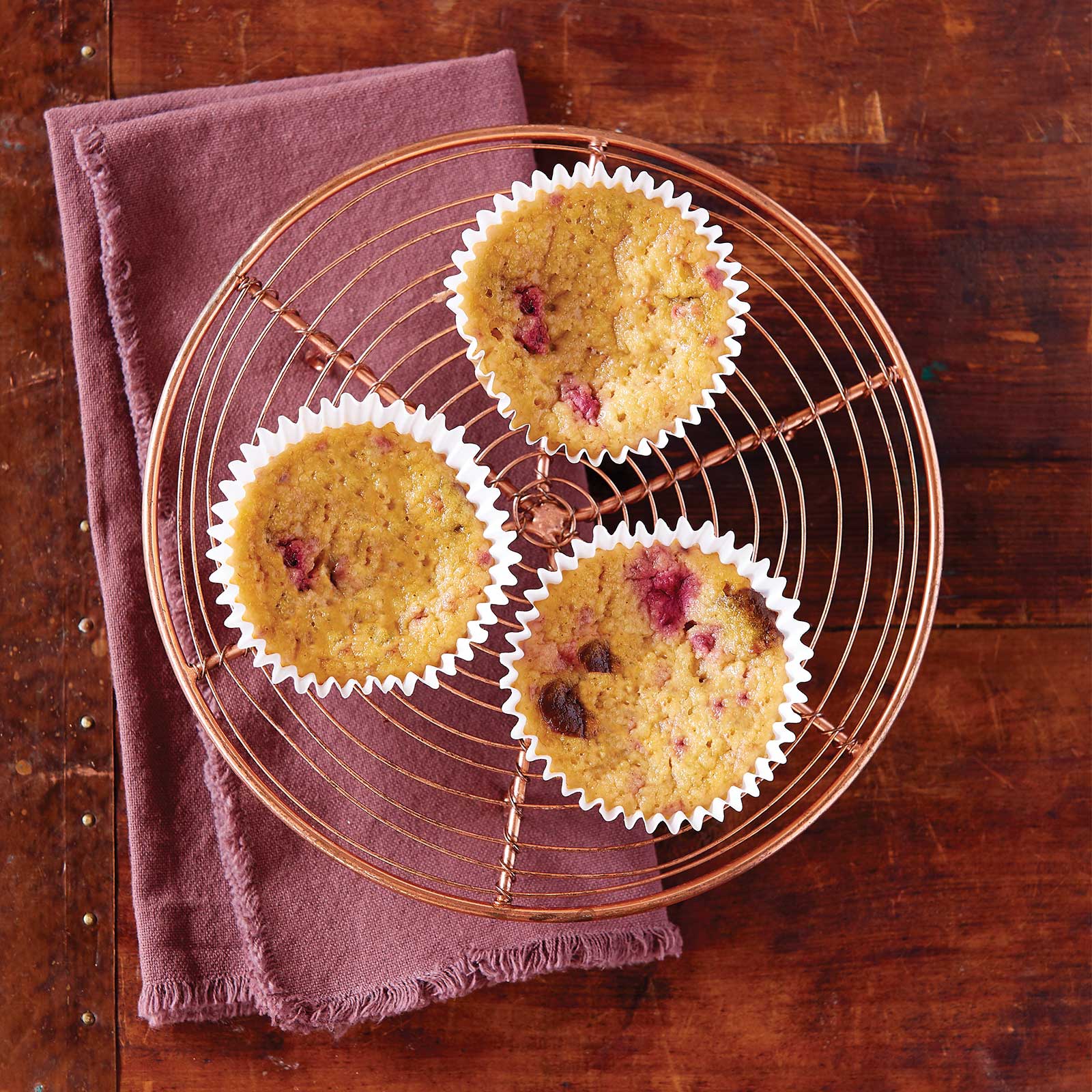 Three grain-free raspberry chocolate muffins sitting on a round wire cooling tray. Shot overhead. The cooling tray is sitting on a mauve napkin.