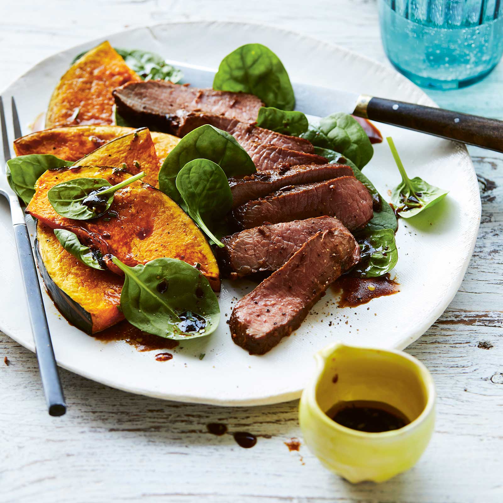 White plate, with slices of lamb roast pumpkin wedges and fresh spinach leaves drizzled with a balsamic dressing.