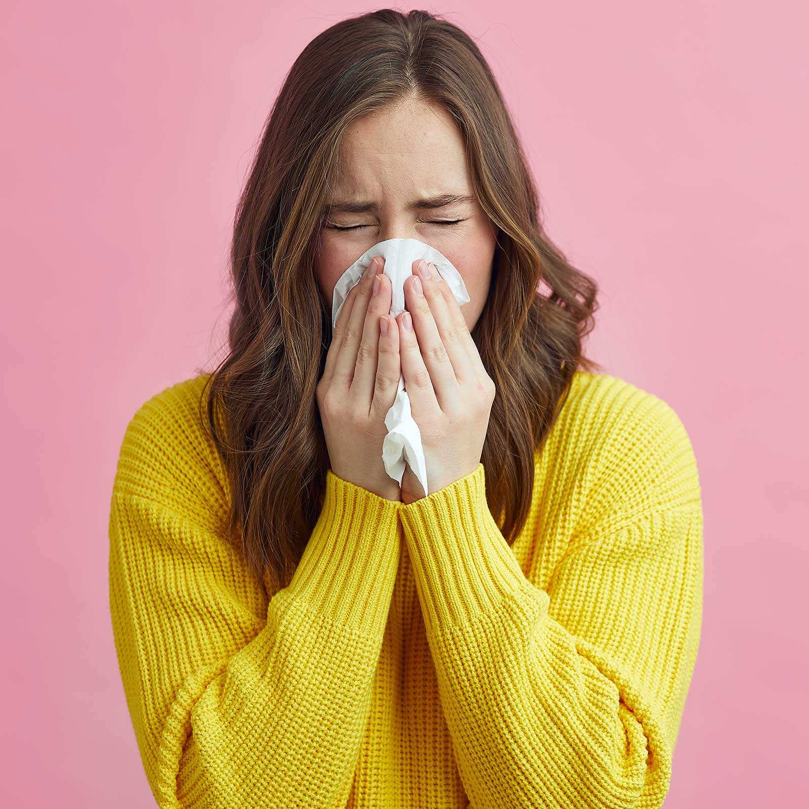 Woman in yellow jumper sneezing. Covers mouth and nose with a tissue.