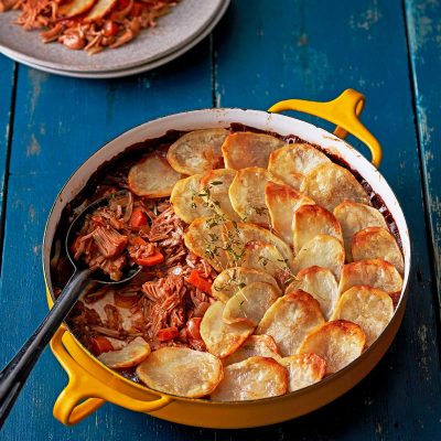 Vegan Lancashire Hotpot in a yellow baking dish with handles. A scoop has been removed and sits on a plate at the back. A spoon is in the hotpot ready to serve more. Slice potato is golden on top of the hotpot.
