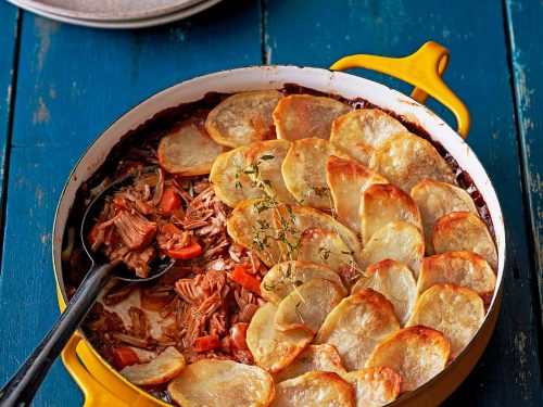 Vegan Lancashire Hotpot in a yellow baking dish with handles. A scoop has been removed and sits on a plate at the back. A spoon is in the hotpot ready to serve more. Slice potato is golden on top of the hotpot.