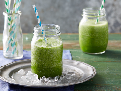 Two glass jars filled with a green smoothie. Both glasses have green and white striped straws. One glass jar site on a silver plate filled with ice.