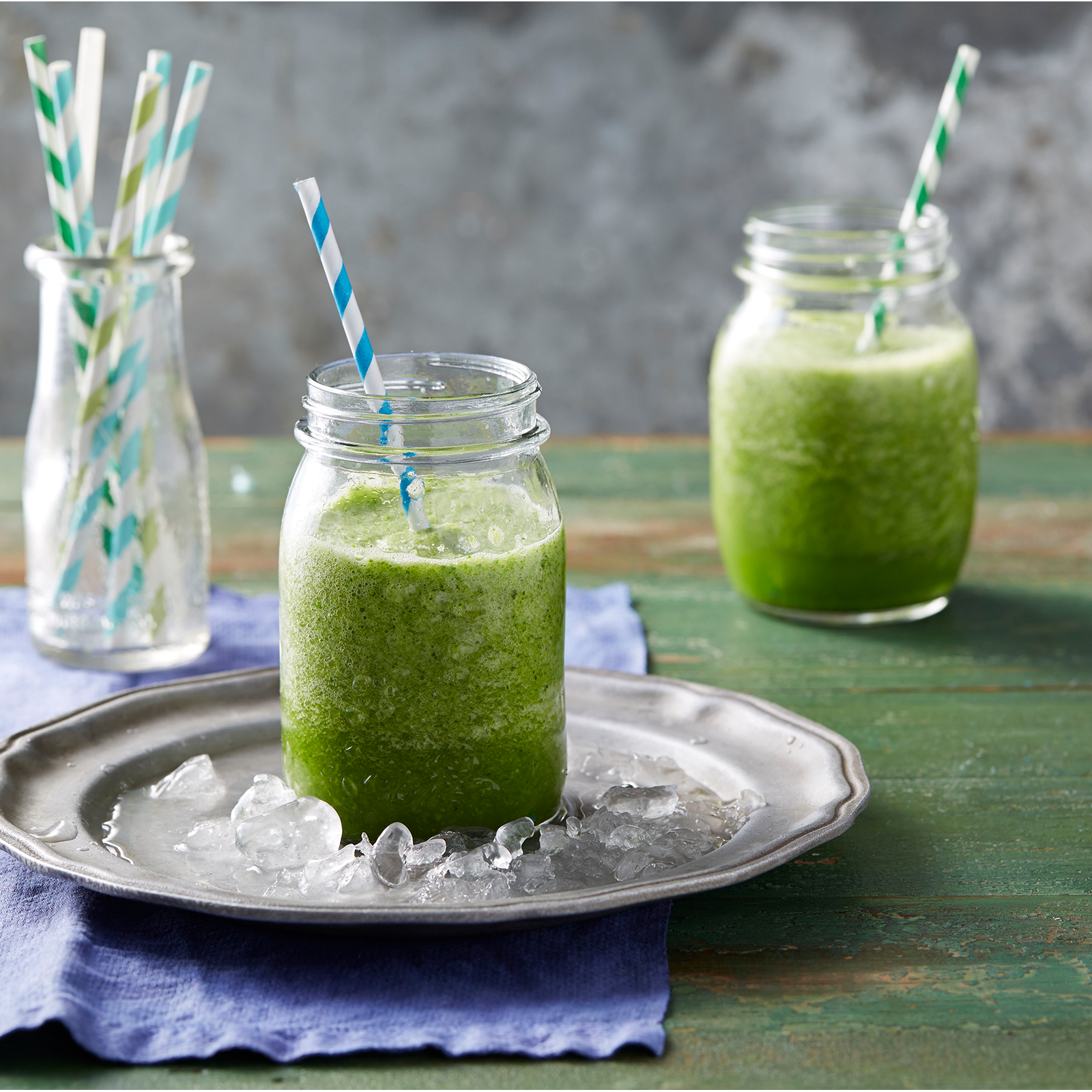 Two glass jars filled with a green smoothie. Both glasses have green and white striped straws. One glass jar site on a silver plate filled with ice.