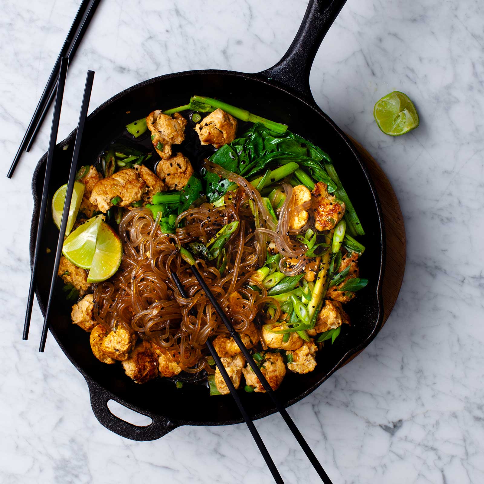 A cast iron pan with gluten-free sweet potato noodles and red curry chicken meatballs. Fresh lime wedges rest on top along with chopsticks, ready to eat straight from the pan.
