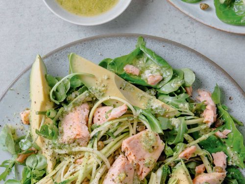 A light blue bowl with the best salon avocado salad including spiralised zucchini, large slices of avocado and chunks of poached salad. At the top of the image is a small bowl of vinaigrette ready for drressing.