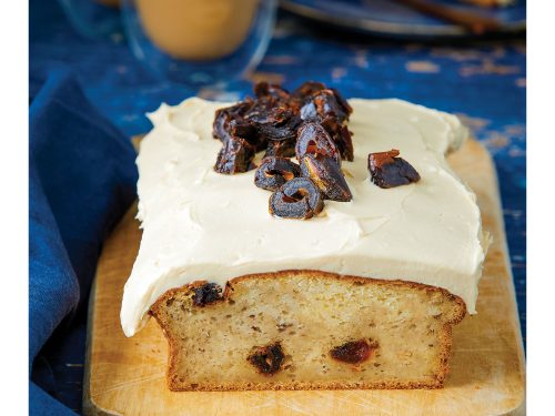 Vegan Tahini and Date Banana Bread with frosting sits on a wooden board. A slice of this gluten-free and vegan banana bread is on a plate next to a coffee at the back.