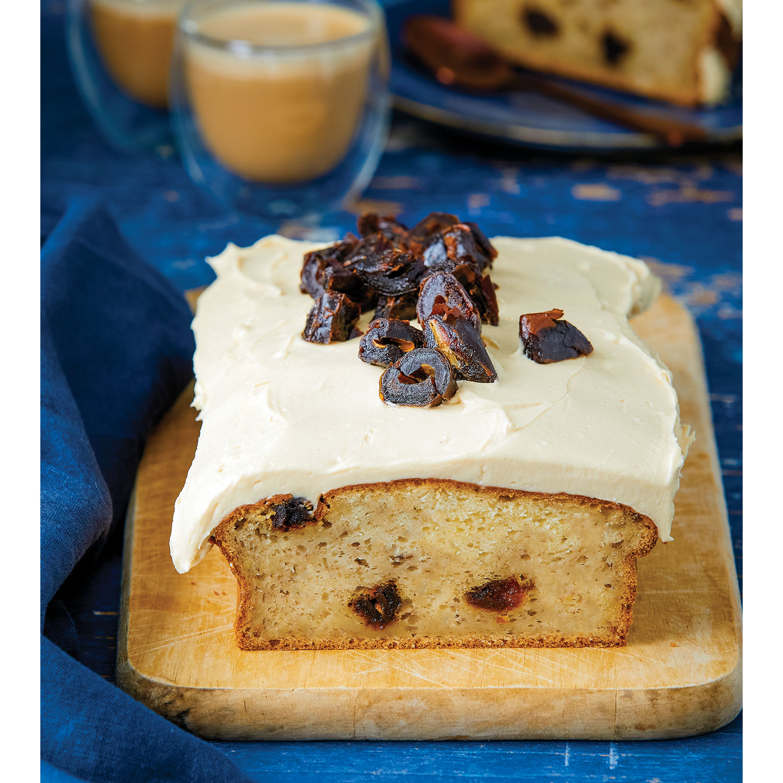 Vegan Tahini and Date Banana Bread with frosting sits on a wooden board. A slice of this gluten-free and vegan banana bread is on a plate next to a coffee at the back.