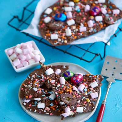Gluten-free easter chocolate bark broken into pieces and arranged on a small round plate. A cake lifter rests next to the plate. At the bake is a wire cooling rack lined with baking paper and more chocolate bark.