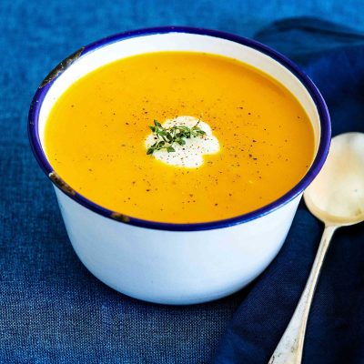 An enamel bowl is filled with low fodmap curried vegetable soup. A dollop of coconut cream sits on top and a soup spoon rests to the right-hand side.