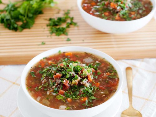 A white bowl rest on a plate. It's filled with mixed lentil quinoa soup and topped with fresh herbs. A gold soup spoon rests to the side. On a wooden board at the back is a bunch of fresh parsley and another white bowl filled with mixed lentil quinoa soup.