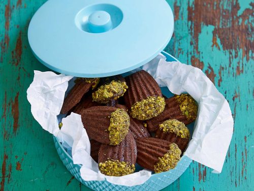 A vintage light blue biscuit container is lined with baking paper and filled with gluten-free mocha madelines that have the tops dipped in chocolate and crushed pistachios. The lid to the container rests on the edge.
