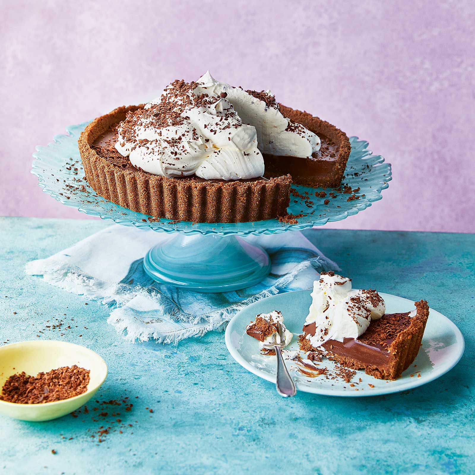 Gluten-Free No Bake Chocolate Cream Pie sits on a blue cake stand. It's topped with cream and crated chocolate. A slice has been remove and sits on a plate in front.