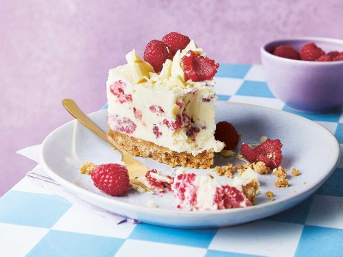 A slice of white chocolate no-bake cheese cake it on a plate, half of the cheesecake has been eaten and there are crumbs and a fork on the plate. The plate rests on a table with a blue and white check table cloth. Next to the plate is a purple bowl with fresh raspberries.