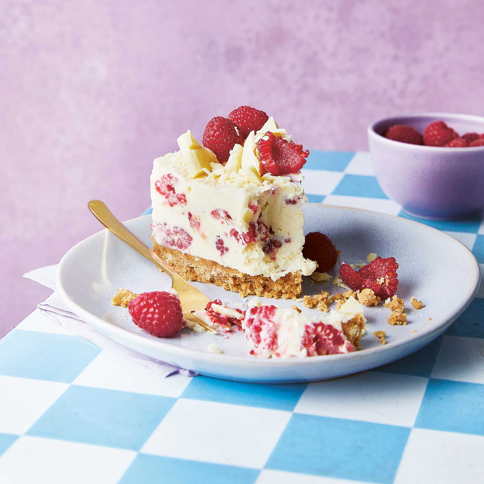 A slice of white chocolate no-bake cheese cake it on a plate, half of the cheesecake has been eaten and there are crumbs and a fork on the plate. The plate rests on a table with a blue and white check table cloth. Next to the plate is a purple bowl with fresh raspberries.
