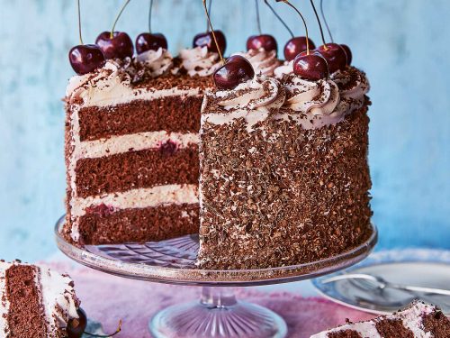 A gluten-free black forest gateau is on a glass cake stand. Two slices have been removed and they rest on small plates in front of the cake. Fresh cherries decorate the top of the black forest gateau.