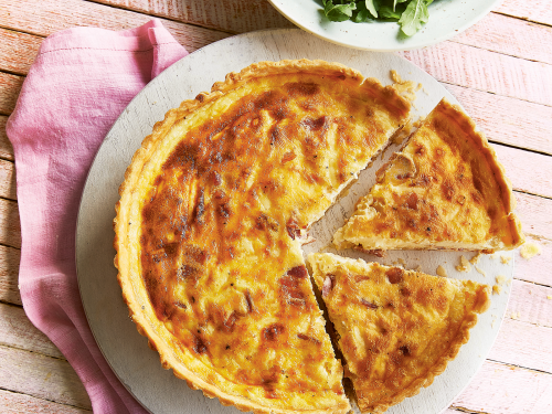 Becky Excell's gluten-free quiche lorraine on a flat round plate. Two slices of quiche have been cut ready to serve. The plate rests on a pink napkin and a bowl of green salad is at the back.