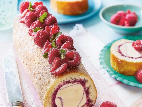 A gluten-free raspberry ripple arctic roll decorated with fresh raspberries and mint leaves is resting on a pink chopping board. Two slices have been placed on brighly coloured plates to the side and back of the chopping board.