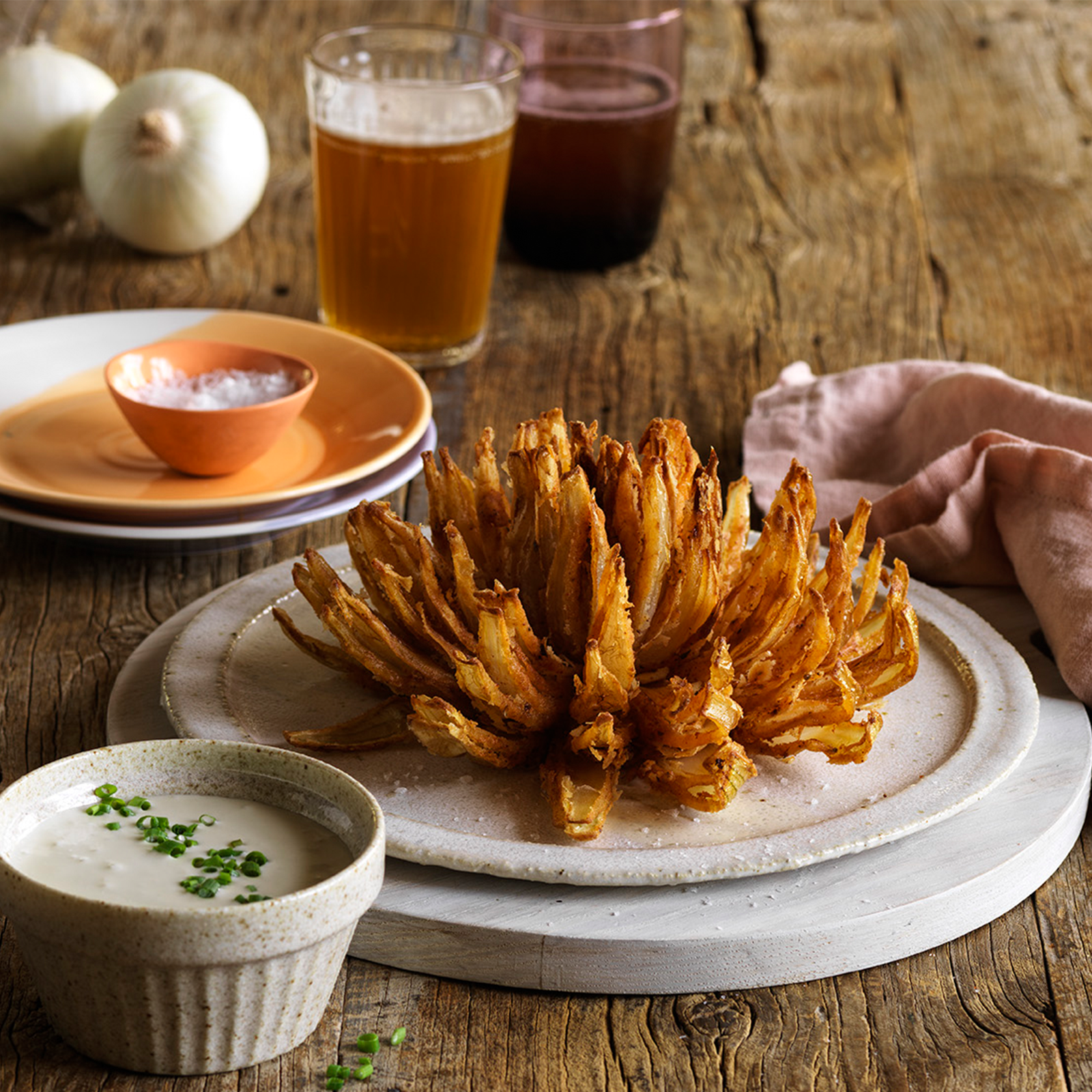 A blooming onion sits on a white plate. In front of the plate is a small bowl with blue cheese tip garnished with fresh herbs. A schooner of gluten-free beer sits at the back.