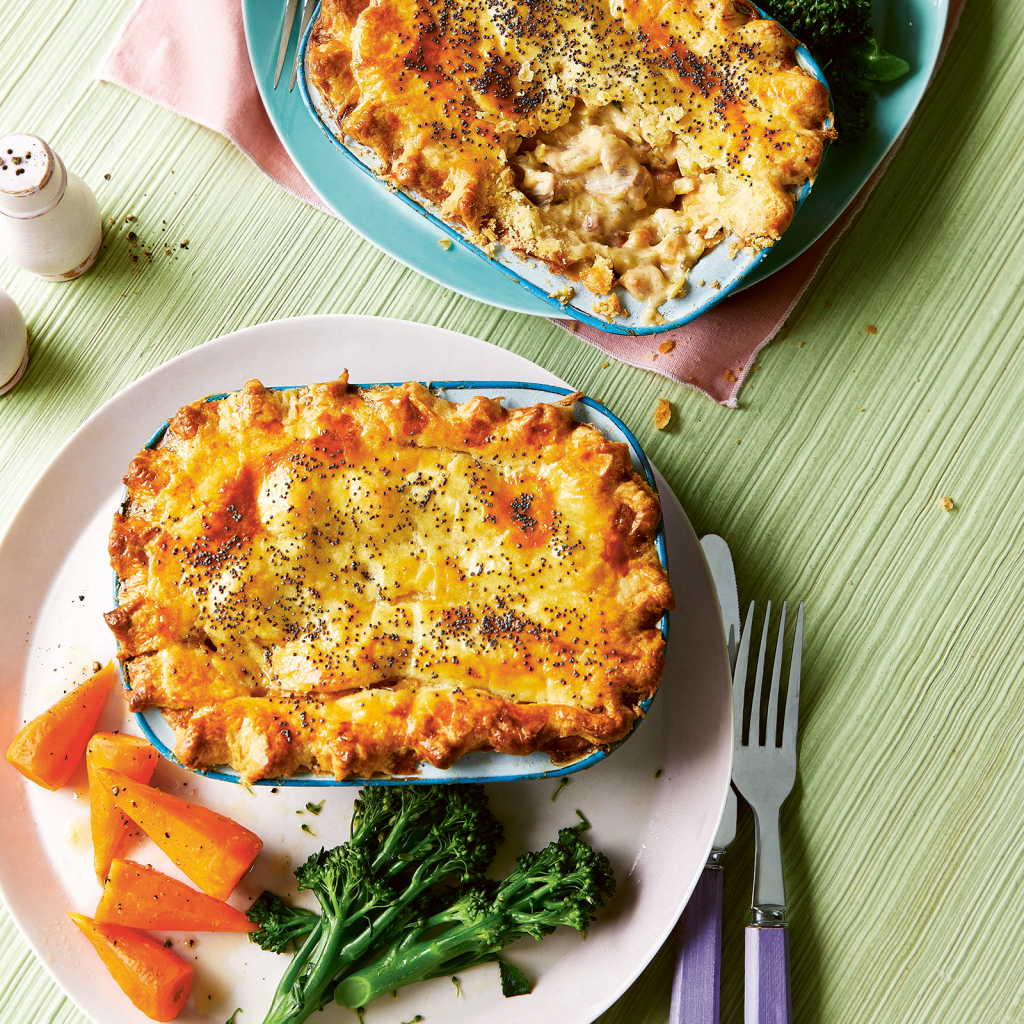 An overhead image featuring two mini chicken bacon and leek pies in individual pie dishes placed on a dinner plate with steamed carrot and broccolini.