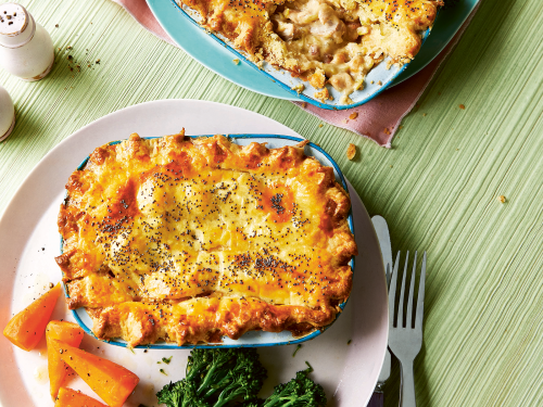 An overhead image featuring two mini chicken bacon and leek pies in individual pie dishes placed on a dinner plate with steamed carrot and broccolini.
