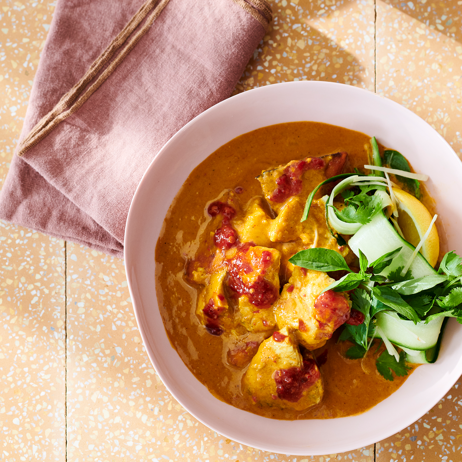 A pink bowl is filled with coconut fish curry and garnished with fresh herbs and thinly sliced cucumber. A dusty pink napkin rests to the side.