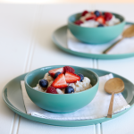 Two green/blue bowls of vegan coconut rice each sit on a large flat plate with a napkin and gold spoon. The rice is topped with fresh strawberries and blueberries.
