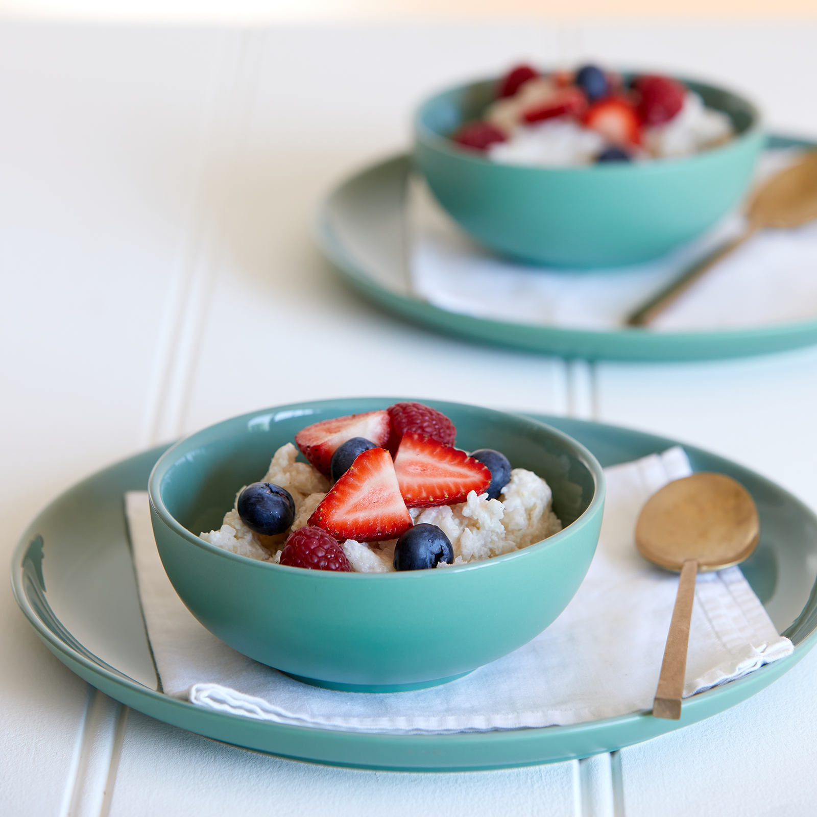 Two green/blue bowls of vegan coconut rice each sit on a large flat plate with a napkin and gold spoon. The rice is topped with fresh strawberries and blueberries.