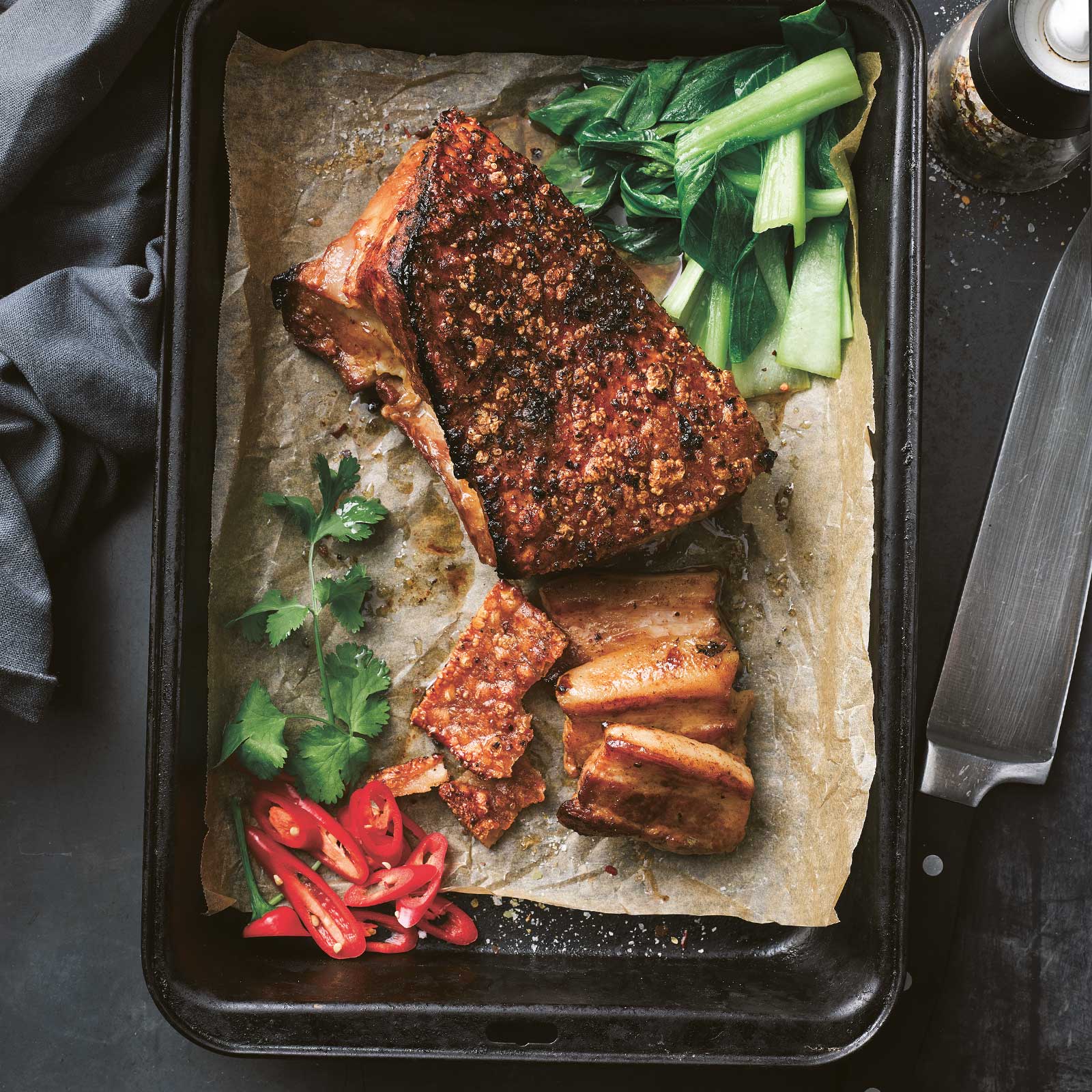 Crispy roast pork belly sits on a roasting pan linked with baking paper. Fresh chilli and herbs decorate.
