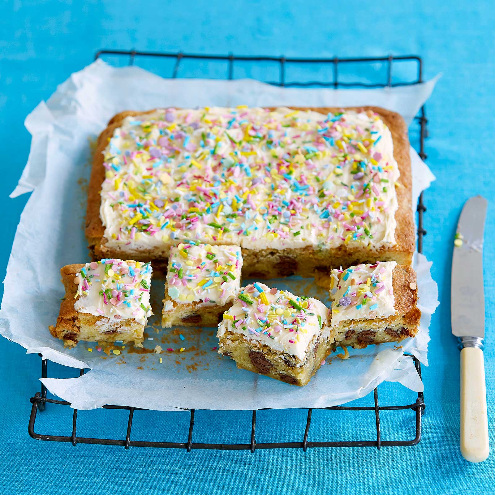 Double-choc blondies with white icing and sprinkles sitting on baking paper on a wire cooling rack. A row of blondies have been sliced into squares and a knife is on the right hand side ready to slice more.
