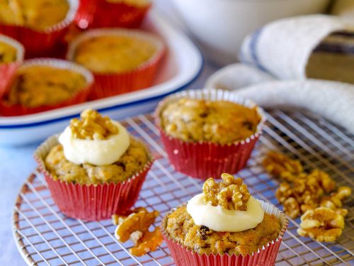 Three egg-free banana walnut muffins sit on a wire cooling rack. Two are iced with cream-cheese frosting and walnut pieces. At the back an enamel dish is full of muffins waiting to be iced.