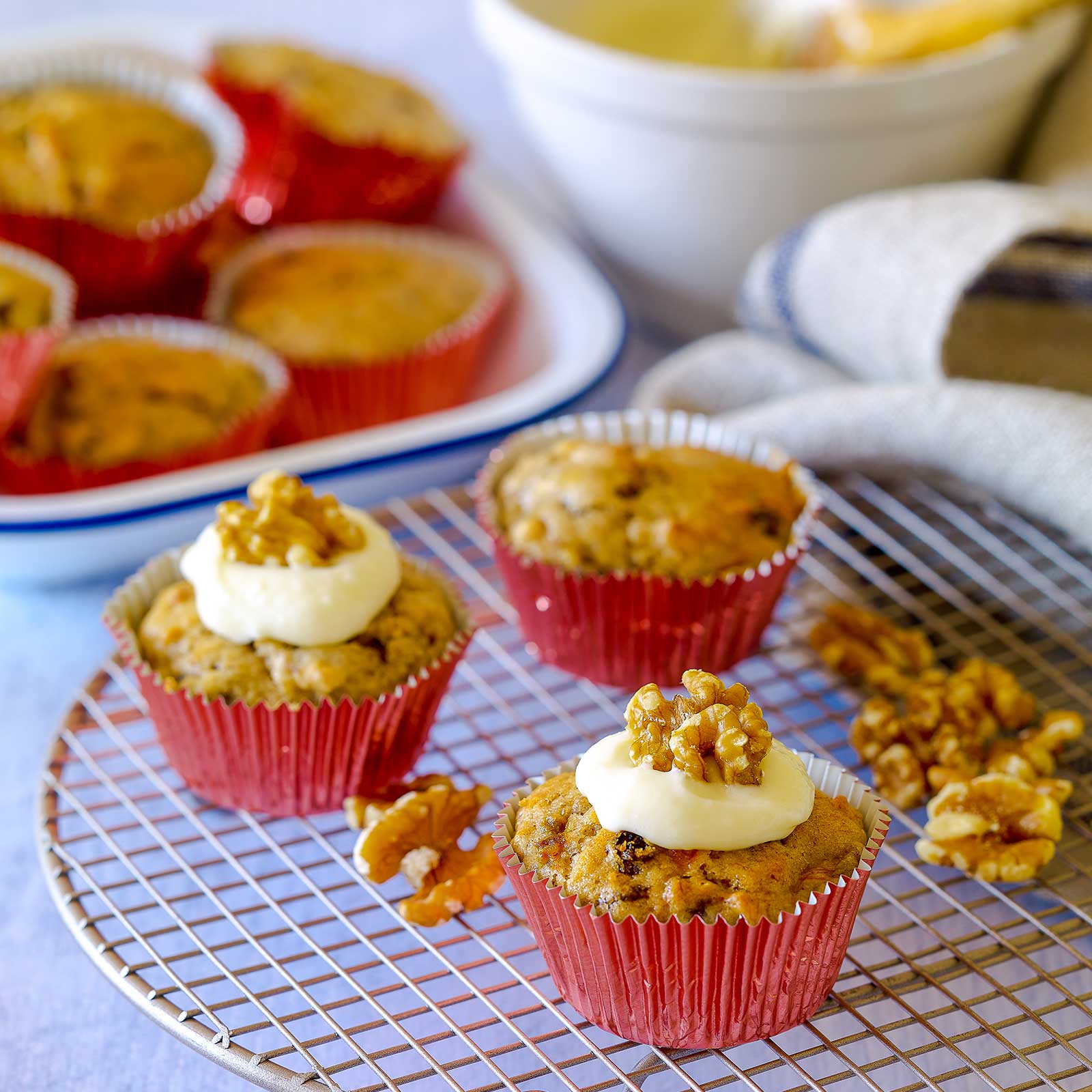 Three egg-free banana walnut muffins sit on a wire cooling rack. Two are iced with cream-cheese frosting and walnut pieces. At the back an enamel dish is full of muffins waiting to be iced.