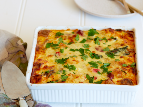 A large white baking dish with gluten-free breakfast strata topped with fresh herbs. A cake lift sits to the side ready to serve onto plates.