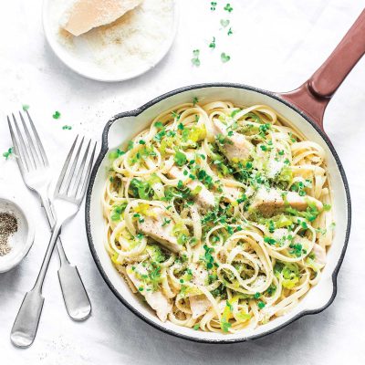 A white saucepan with wooden handle with gluten-free chicken & leek fettucine. The pasta has been garnished with micro herbs and grated Parmesan.