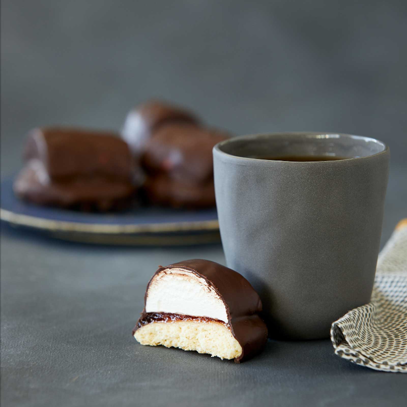 Half a gluten-free chocolate royal biscuit sits in front of a fine ceramic grey mug. Behind the mug is a grey plate with more choc-royals waiting to be served.