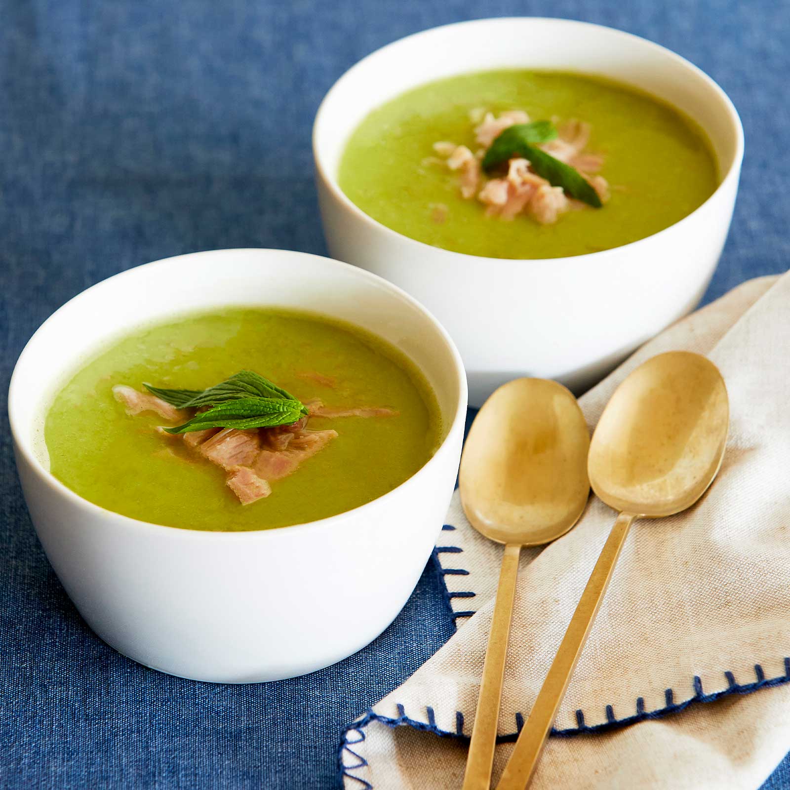 Two white bowls filled with gluten-free fresh pea & ham soup. Topped with extra pieces of ham and mint leaves. Two gold soup spoons rest on a cream napkin to the right of the bowls.