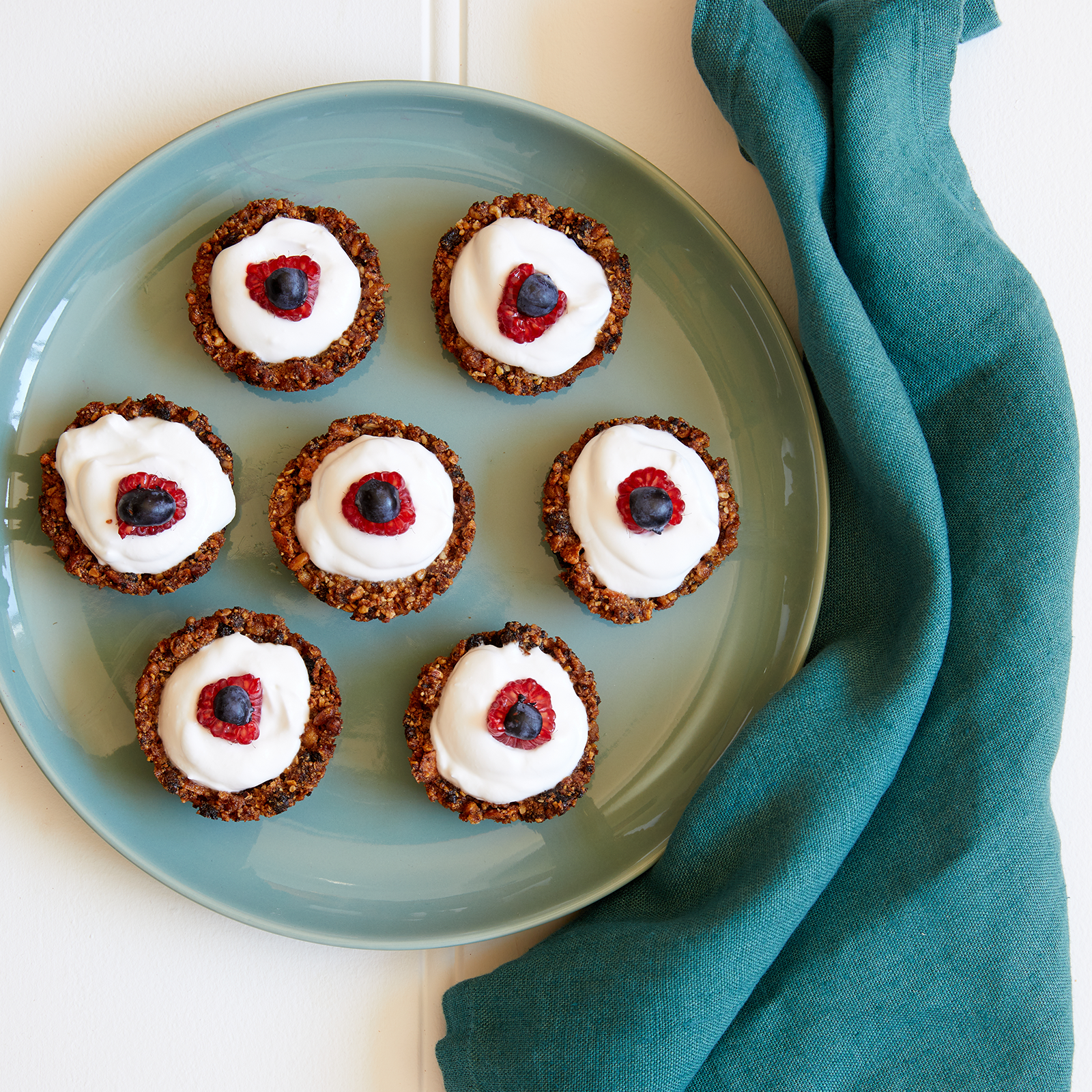 Overhead shot of individual gluten-free granola tarts filled with vegan yoghurt and topped with berries. The tarts are sitting on a large green plate on a white background.