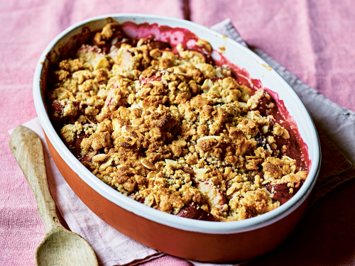 An oval baking dish filled with gluten-free plum crumble. The dish rests on pink fabric with a wooden serving spoon on the side.