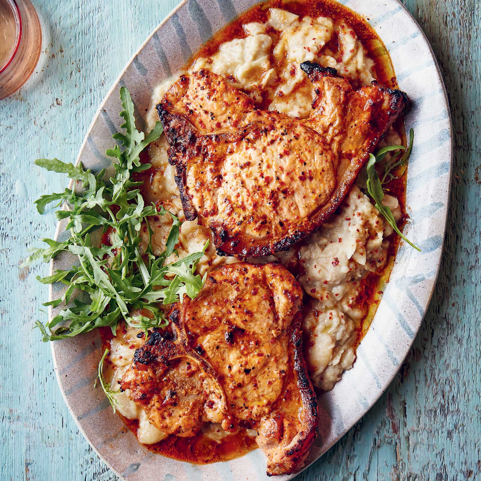 Two pork chops are on a blue and white platter sitting on top of butter bean mash. Fresh rockers sits to the side as a garnish.