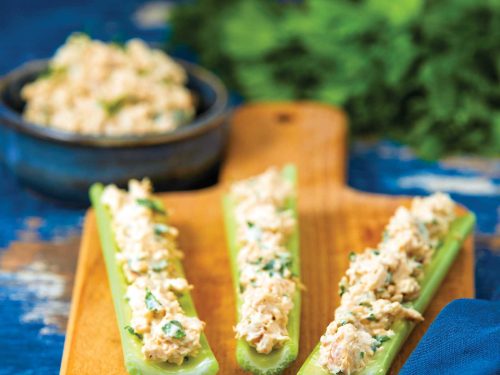Three pieces of celery rest on a wooden chopping board. The celery is filled with spicy chicken and topped with chopped parsley. A bowl of more spicy chicken rests at the back next to a bunch of flat-leaf parsley.