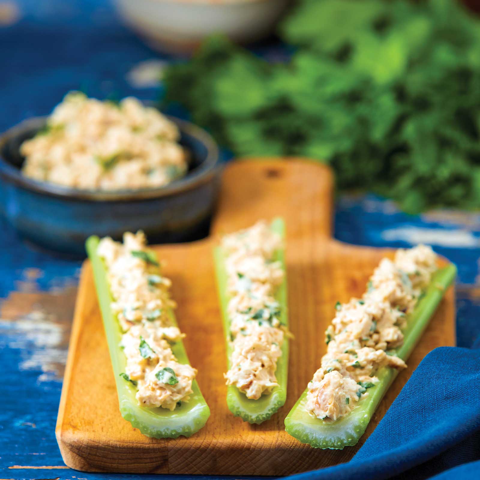 Three pieces of celery rest on a wooden chopping board. The celery is filled with spicy chicken and topped with chopped parsley. A bowl of more spicy chicken rests at the back next to a bunch of flat-leaf parsley.