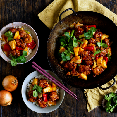 A wok with gluten-free sweet & sour pork sits on lime green fabric. Two bowls with rice and sweet & sour pork rest to the side. A set of chopsticks sit on the side of one bowl.