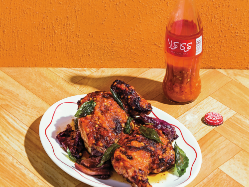 A platter of the best peri peri chicken sits on a wooden bench. The back wall is bright orange and a bottle of chilli sauce sits behind the chicken.