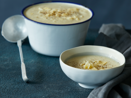 A large enamel bowl with blue rim is at the back of the image filled with the ultimate cheesy cauliflower soup. A ceramic bowl is at the front with a single serve. Both bowls of soup have been topped with shaved Parmesan and cracked parmesan.