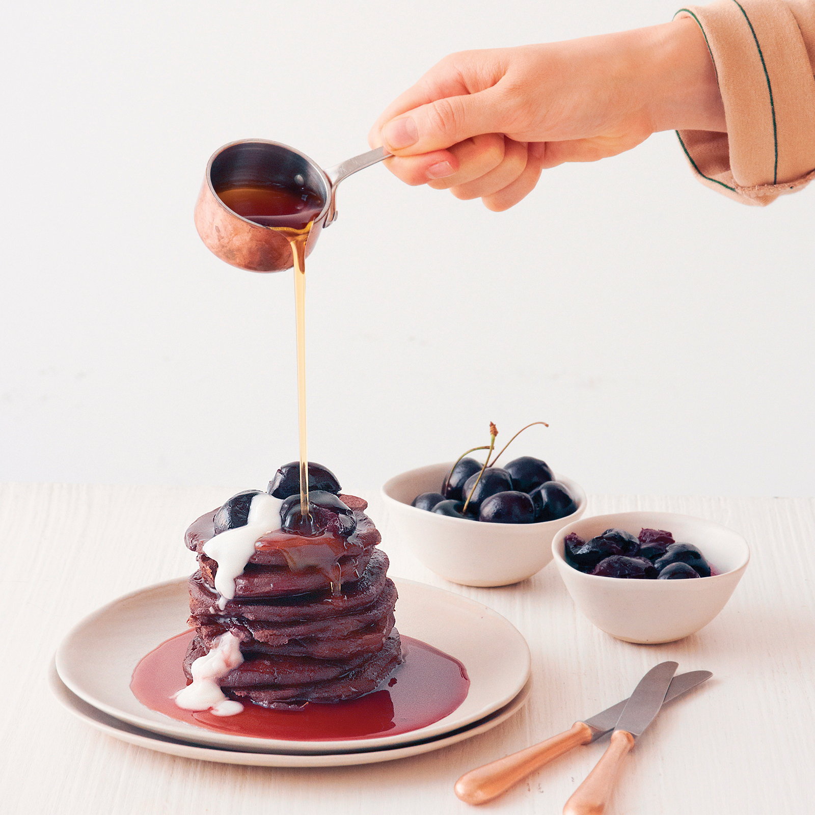 A white plate with a stack of cherry cacao teff pancakes. A hand is over the stack pouring syrup onto the pancakes.