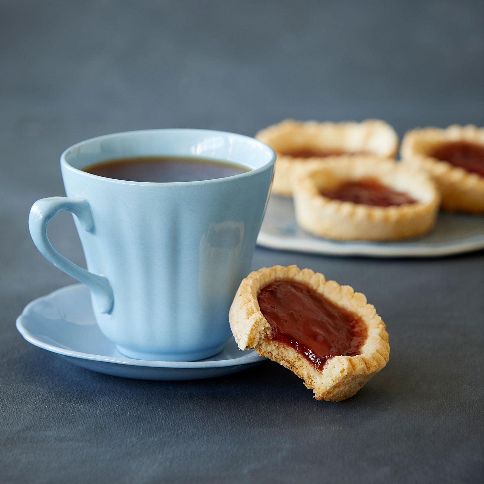 A cup of coffee with a gluten-free jam tart resting on a saucer. At the back a plate contains three more jam tarts.