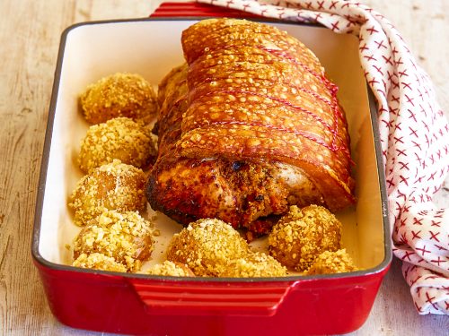 Roast port with gluten-free stuffing sit in a red baking dish surrounded by crispy baked potatoes.