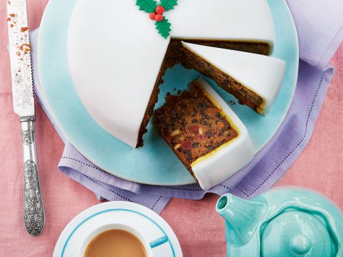 Gluten-Free Christmas Cake by Becky Excell. The cake is covered in white marizpan with fontant holly on top. A segment of the cake is cut so you can see the dried fruit and nuts inside.