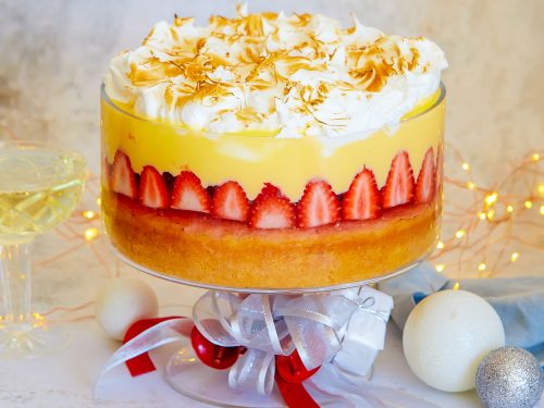 A layered summery strawberry gin trifle is in a glass trifle bowl. The trifle is topped with Italian meringue. Red and silver ribbons have been tied around the stem of the bowl and Christmas decorations and lights are in the background to create a festive scene.