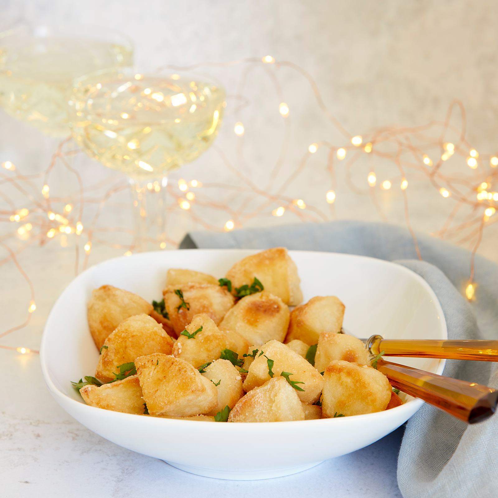Gluten-free crispy baked potatoes in a white bowl with serving utensils ready to serve.The potatoes have been sprinkled with freshly chopped herbs. Fairy lights and champagne glasses are in the background to create a festive feel.